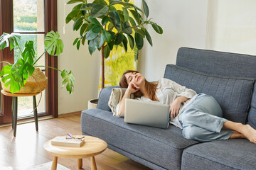 A woman comfortably works from home on a couch, using her laptop and talking on the phone,...