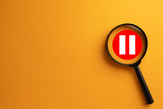 Magnifying glass focusing on a red pause button icon with white bars. Concept of taking a break, stopping for reflection,work hiatus, analyzing an interruption,or media player control.