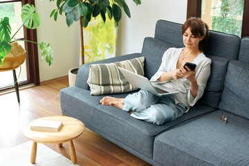 Obraz premium A woman comfortably works from home on a couch, using her laptop and talking on the phone, surrounded by plants and natural light.