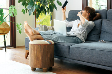 A woman comfortably works from home on a couch, using her laptop and talking on the phone,...
