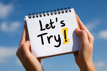 Hands hold notepad with text "Let's Try" against bright blue sky, showcasing motivation and encouragement for new experiences or challenges