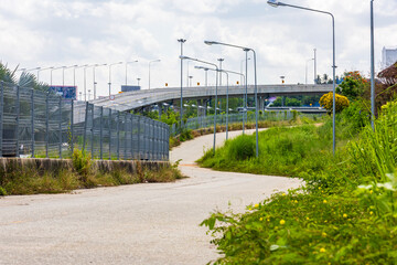 Urban elevated highway and small rural road with overgrown grass, street lights, and fence in foreground under bright blue sky. Transportation infrastructure in suburban area.