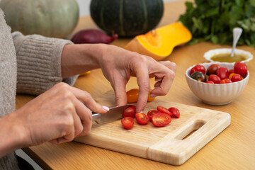 Female hands slicing cherry tomatoes on wooden cutting board with vegetables