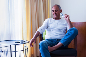 Thoughtful senior man sitting in chair with reading glasses in hand