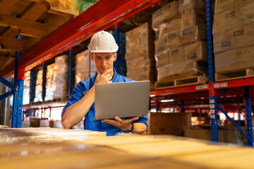 warehouse manager in hard hat using a laptop to analyze inventory data among stacked pallets in a high-bay storage facility.