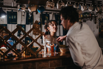 Two people are shown at a warmly lit bar featuring hanging mugs and reflective glasses, conveying a relaxing and conversational atmosphere.