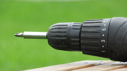 Close-up view of a black electric drill resting on a wooden surface in a green outdoor setting during daylight