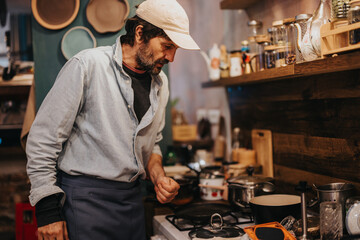 A man preparing food in a warmly lit, rustic-style kitchen. Ingredients, pots, and utensils are displayed, creating a homely and inviting culinary scene.