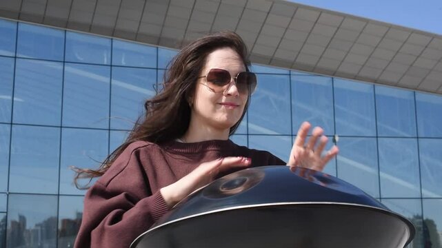 young caucasian woman playing black handpan with hands, ethnic metal drum instrument, sits on stone staircase, scyscraper and business center in background, beautiful relaxing music for meditation