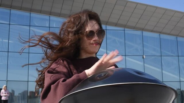 young caucasian woman playing black handpan with hands, ethnic metal drum instrument, sits on stone staircase, scyscraper and business center in background, beautiful relaxing music for meditation