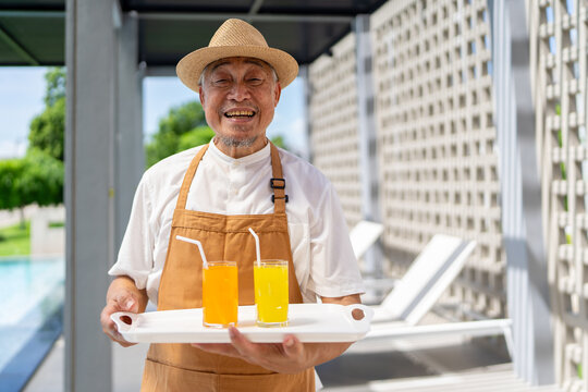 Portrait of cheerful grey-haired man wearing apron uniform serving beverage fruit juice mocktail by swimming pool, asian elderly people do part time job enjoy summer vacation joyful retirement.