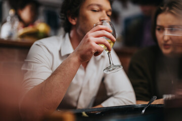 A young man in a white shirt enjoys a glass of wine while sitting indoors, accompanied by friends, in a cozy bar setting. The scene conveys a warm, social, and friendly atmosphere.