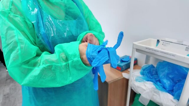 Medical professional putting on blue disposable gloves for safety and hygiene in a healthcare setting, near a cart of individual protective equipment