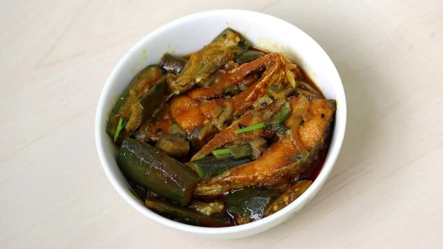 Authentic Bengali brinjal and Hilsa fish curry. Delicious, popular dish served in white bowl on light wood.