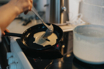 A person pours batter into a skillet on a stove to prepare pancakes. The kitchen setup suggests cooking or baking activity. The image conveys culinary creativity and warm home vibes.