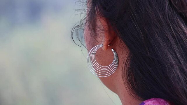 Close-up of Bengali woman's ear with stainless steel hoop earrings. Black hair in breeze, blurred nature background.