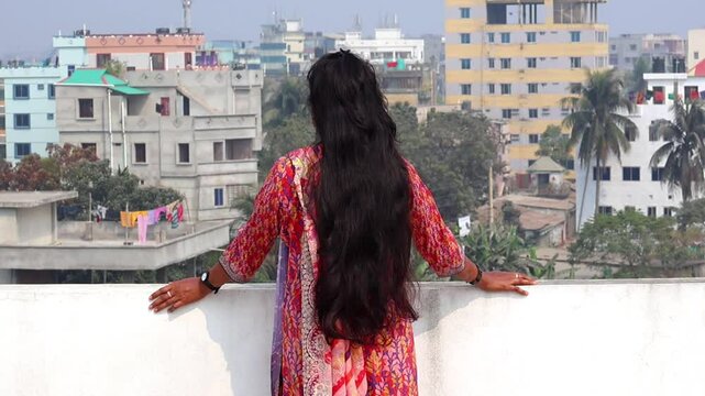 Beautiful Bengali woman in colorful salwar kameez with open black hair, enjoying rooftop morning sunlight and scenic view.
