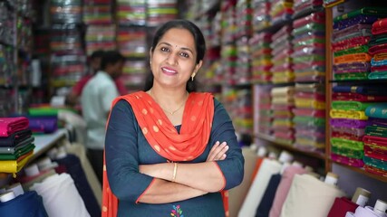 smiling indian senior woman fabric vendor standing with arms crossed in textile shop. small business owner from india in marketplace selling colorful fabrics for clothing material.	

