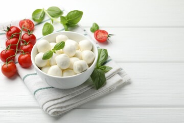 Tasty mozzarella cheese balls in bowl, basil and tomatoes on white wooden table, space for text