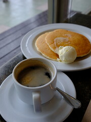 Vertical shot of hot coffee and pancakes with butter on table by the window. Cozy breakfast scene with warm tones, perfect for food blogs, lifestyle content, remote work, and editorial visuals.