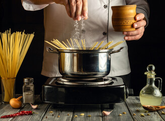 Chef is pouring salt into a pot of boiling water while holding a wooden cup. Spaghetti is placed in...