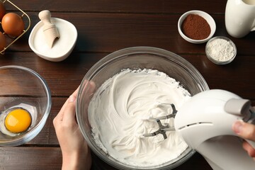 Woman whisking cream with hand mixer at wooden table, top view