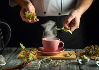 Hands are seen adding fresh herbal leaves to a steaming cup of tea on a wooden table. Surrounding...