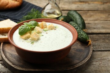Tasty cucumber soup with croutons, basil, bread and vegetables on wooden table, closeup