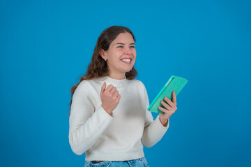 Happy young woman in a white sweater holding a green tablet and smiling with a fist raised in a gesture of triumph, standing against a solid blue background, symbolizing success and positive results 