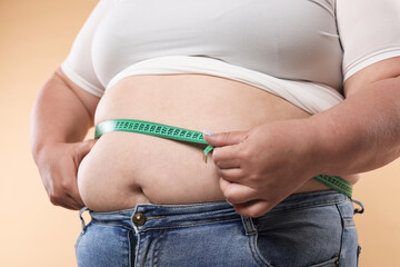 Overweight woman measuring her belly with tape on beige background, closeup