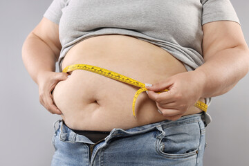 Overweight woman measuring her belly with tape on grey background, closeup