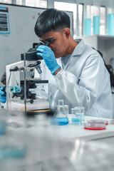 Fototapeta premium Focused Asian male scientist using microscope in modern laboratory with DNA structure chart in background. Concept of biotechnology, genetics research, molecular biology and advanced healthcare.