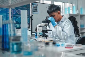 Focused Asian male scientist using microscope in modern laboratory with DNA structure chart in...