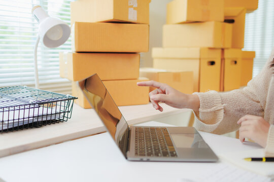 Freelance woman working from home office, packaging orders and pointing at laptop screen while managing online marketing and delivery for her small business