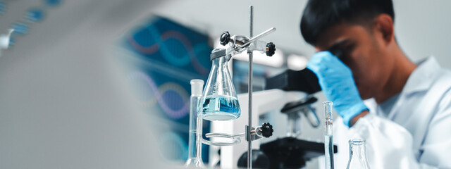 Focused Asian male scientist using microscope in modern laboratory with DNA structure chart in...