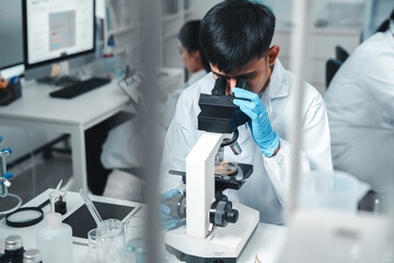 Focused Asian male scientist using microscope in modern laboratory with DNA structure chart in...