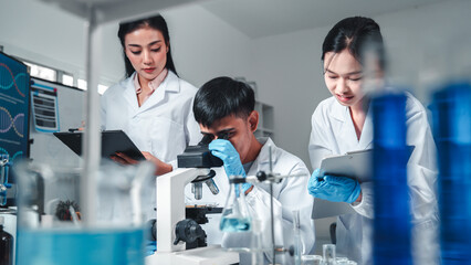 Focused Asian male scientist using microscope in modern laboratory with DNA structure chart in...