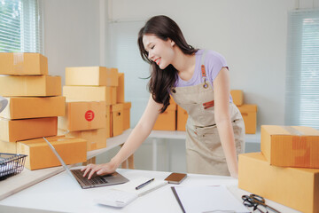 Young entrepreneur managing her online business, using a laptop to check orders and shipments in a warehouse full of cardboard boxes, smiling and focused on her work