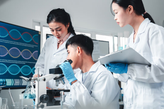 Three young scientists in lab coats collaborate on a research project using a microscope and taking notes, surrounded by lab equipment and blue liquids, showing teamwork and scientific analysis.
