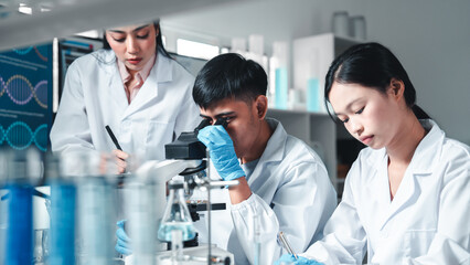 Group of young scientists in lab coats collaborate in a high-tech laboratory, analyzing samples through a microscope and documenting results, symbolizing innovation, teamwork, and modern research.