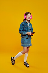 Fototapeta premium A smiling little girl wearing denim and pink headphones, holding a smartphone, enjoying music joyfully. Ideal for technology and lifestyle advertisements.