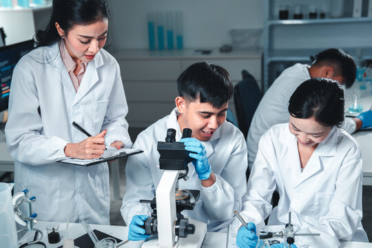 Group of young scientists in lab coats collaborate in a high-tech laboratory, analyzing samples through a microscope and documenting results, symbolizing innovation, teamwork, and modern research. - Powered by Adobe