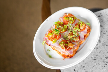 A fresh open-faced sandwich on a white plate, topped with sliced tomatoes, ham, microgreens, and a drizzle of sauce, placed on a speckled countertop. Ideal for food photography