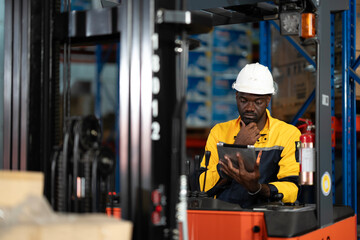 Black forklift driver checks information on a tablet while sitting on the vehicle. Concentrated on digital tools to manage warehouse tasks effectively. © Wanwajee