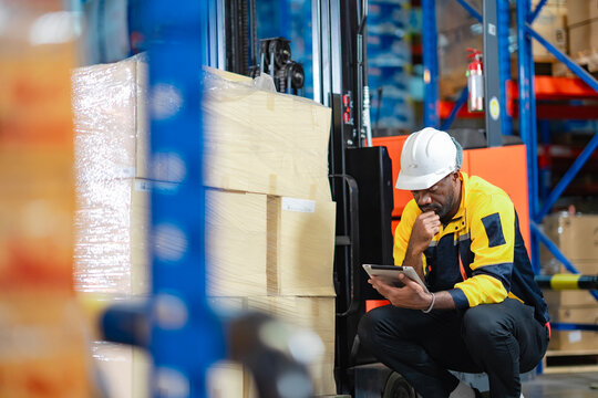 Forklift operator checking inventory in warehouse. Black male worker in uniform and helmet using tablet to review shipment packaging and logistics.