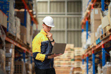 Focused industrial worker stands in a warehouse corridor, updating stock records on his laptop and reviewing shelf contents. Logistic industry with technology...