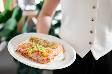 A waiter in white uniform holds a plate with a delicious open-faced sandwich topped with fresh tomato slices, greens, and avocado. The blurred green background creates a fresh and inviting atmosphere