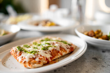 A close-up of a rectangular pizza with melted cheese and green herbs on a white plate, presented in a dining setting with a blurred background. The pizza looks freshly baked and appetizing