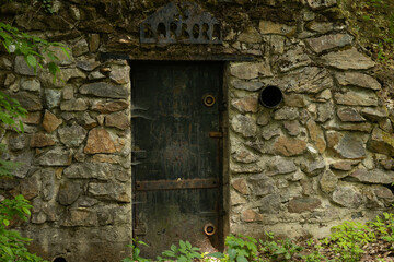 An old stone door set in a weathered wall, surrounded by greenery. The door is dark and rustic, with metal fittings and a small circular vent.
