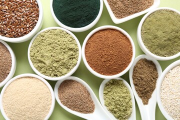 Different superfood powders in bowls and spoons on pale green background, flat lay
